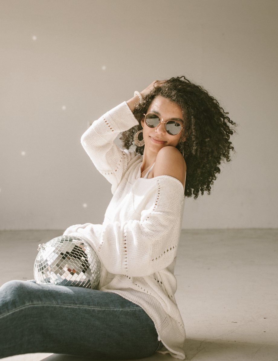Allie smiling in sunglasses and a white sweater, one hand in her curls, sitting beside a disco ball