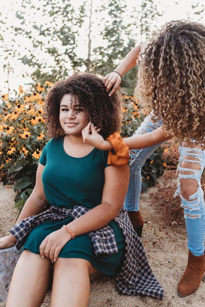 Outdoors among yellow flowers, a stylist fluffs a smiling guest’s full, curly hair